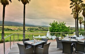 Uma área de refeições ao ar livre com mesas e cadeiras, com vista para uma paisagem deslumbrante com campos e montanhas.