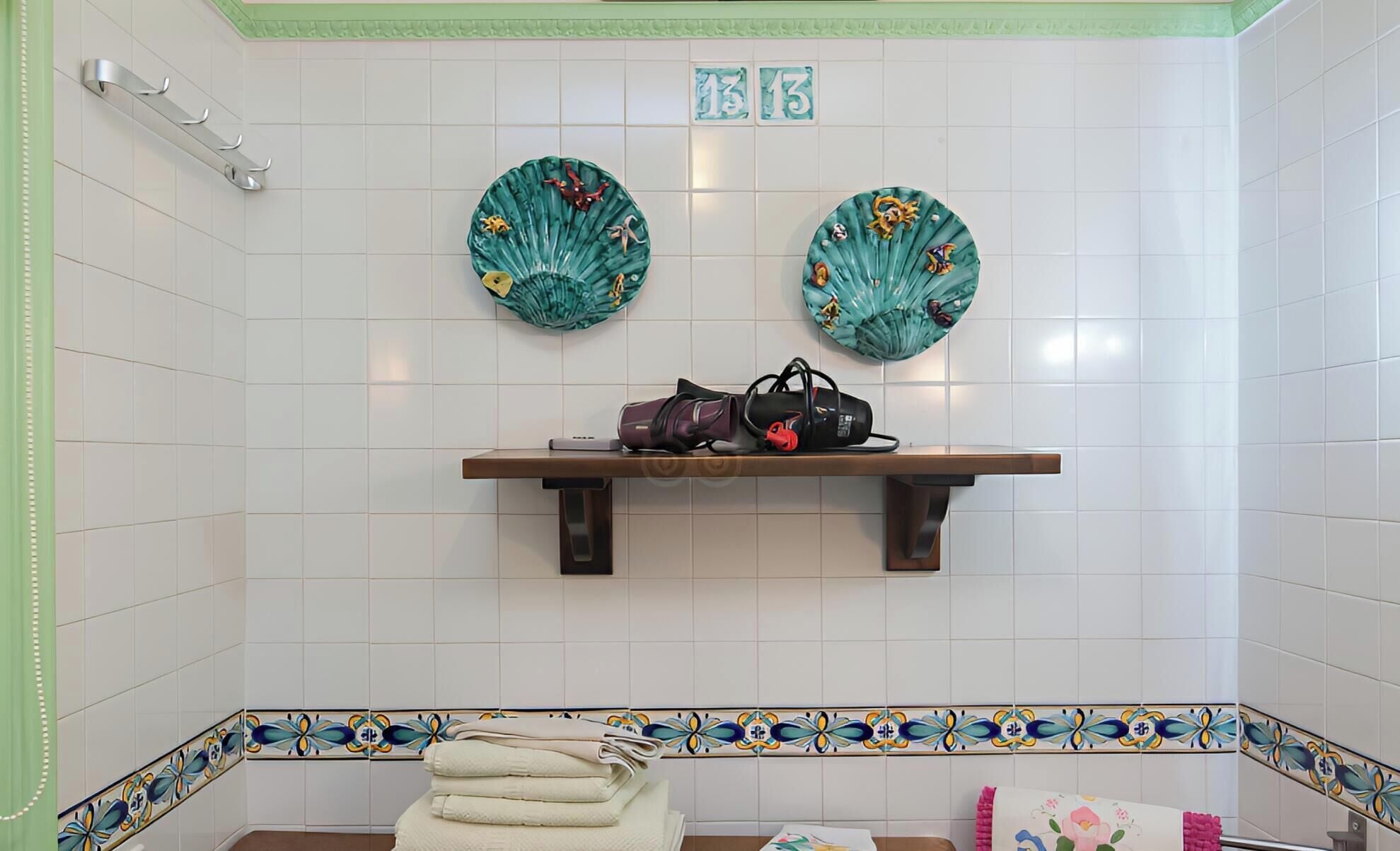Une salle de bain avec des murs carrelés de carreaux blancs, une étagère en bois sur laquelle se trouve un sac, deux éventails décoratifs et une pile de serviettes.