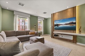 A living room with a sectional sofa, a flat-screen TV mounted on a wooden wall, and a window with blinds. - Last Minute Disney Getaway! Beautiful Townhouse & Private Balcony. Book Now! #172665 (Orlando)