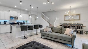 A modern kitchen with white cabinets, a central island with bar stools, a dining area with a wooden table and chairs, and a staircase leading to an upper level. - Last Minute Disney Getaway! Private Pool & Themed Bedrooms. Book Now! #172669 (Orlando)