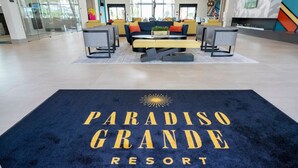 Eine Hotelhalle mit einer Rezeption, einer Sitzecke und einem blauen Teppich mit der Aufschrift 'PARADISO GRANDE RESORT'.