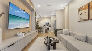 Ein modernes Wohnzimmer mit großem Fernseher, einer Couch, einem Couchtisch und einer Küche im Hintergrund.