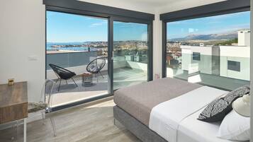 Un dormitorio con una cama grande, una silla y vistas a un paisaje urbano a través de ventanales.
