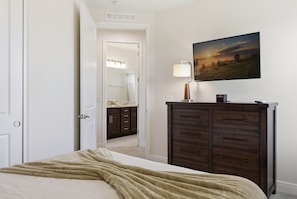 Un dormitorio con cama, cómoda, un cuadro y un baño visible a través de una puerta abierta.