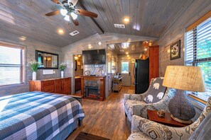 Interior acogedor de una cabaña con cama, sofá, chimenea, zona de cocina y ventilador de techo.