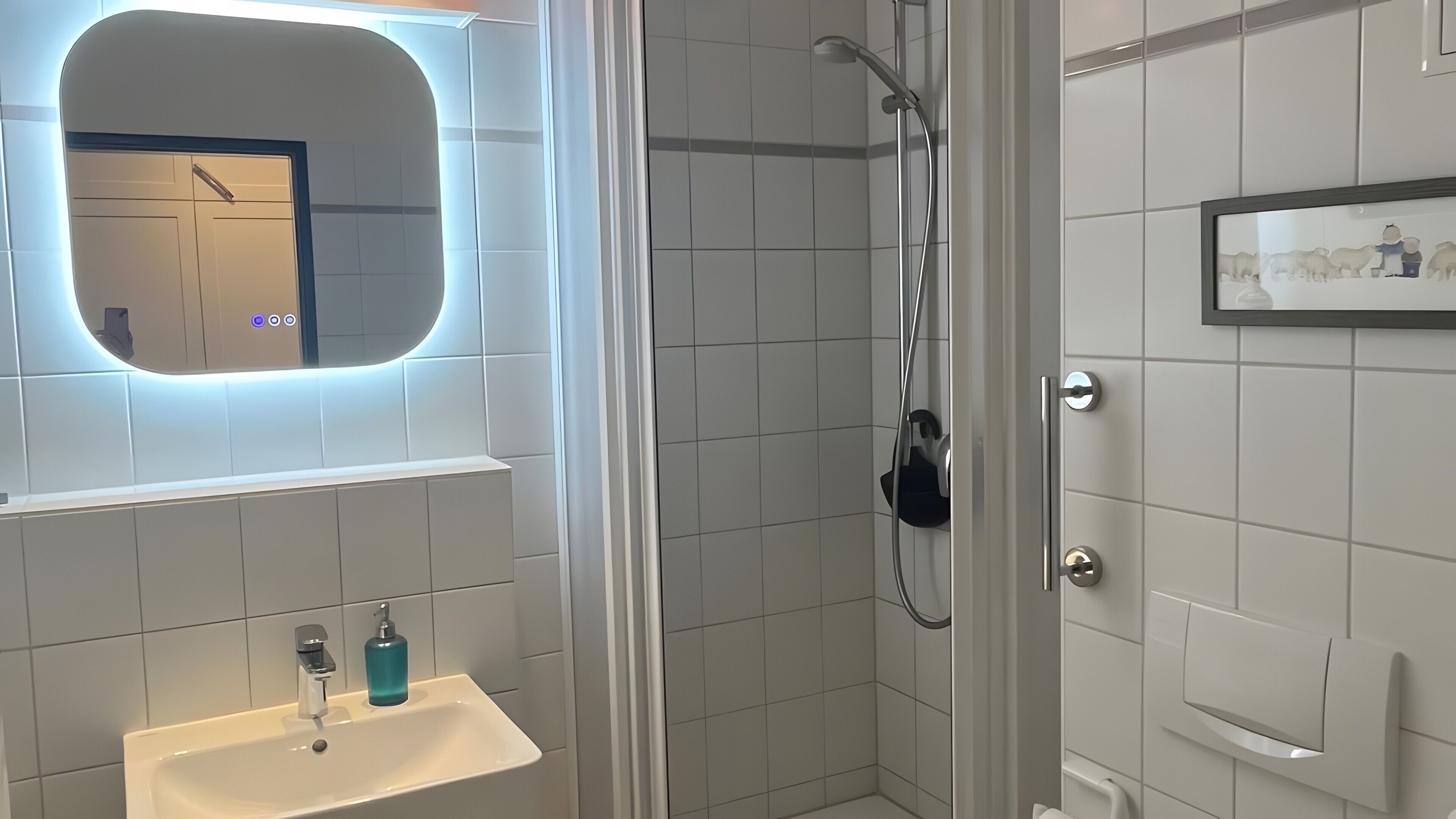 Ein Badezimmer mit einem weißen Waschbecken, einem Spiegel mit Beleuchtung, einer Dusche inklusive Handbrause und einem gerahmten Bild an der Wand.