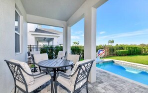 Un espacio al aire libre cubierto con piscina, una mesa con sillas y vistas a una casa al fondo.