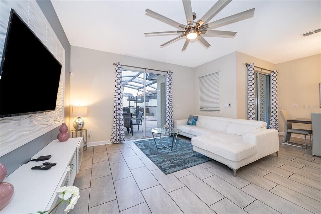 A modern living room with a white sofa, a flat-screen TV, a ceiling fan, and a glass door leading to an outdoor area.