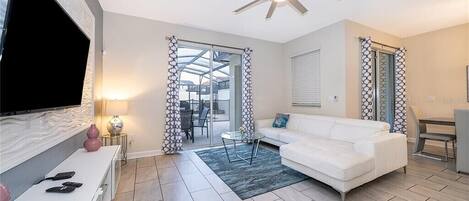 A modern living room with a white sofa, a flat-screen TV, a ceiling fan, and a glass door leading to an outdoor area.