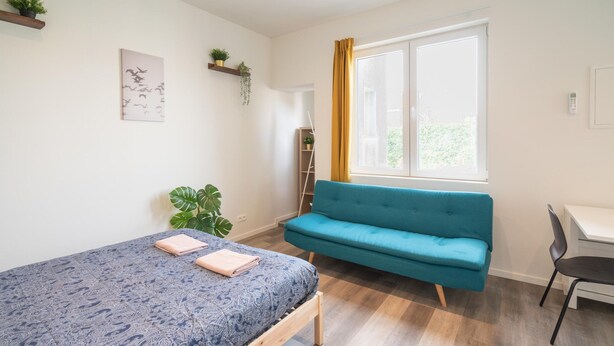 A bedroom with a bed, a blue sofa, a desk, and a window with curtains. - Cosy Apartment in Antwerp Centre (Antwerp)