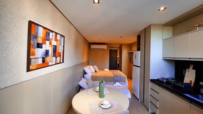 Quarto de hotel compacto com uma cama, uma mesinha com um vaso, um refrigerador e uma cozinha compacta.