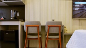 Ein Hotelzimmer mit Küchenzeile, zwei Holzstühlen, einem Bett und einem Fernseher, der eine Speisekarte zeigt.