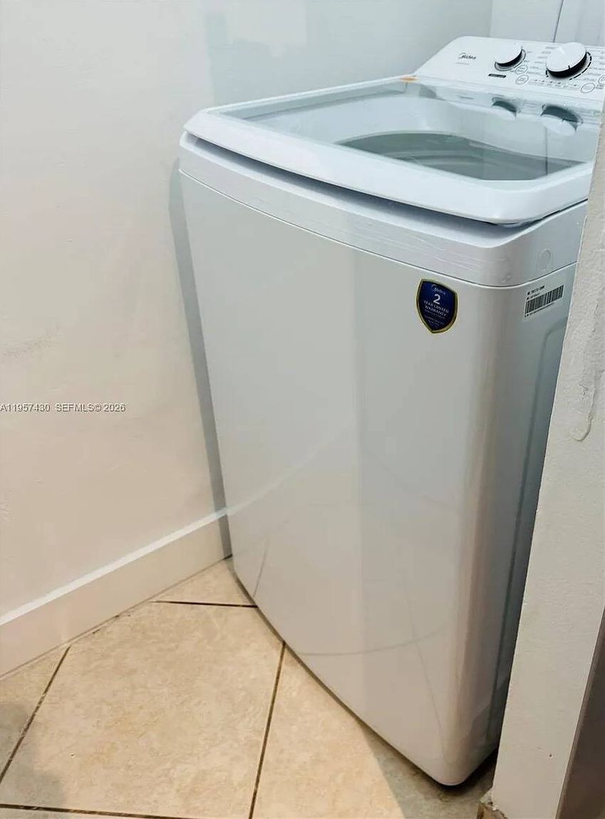 A white front-loading washing machine in a corner of a room with tiled flooring.