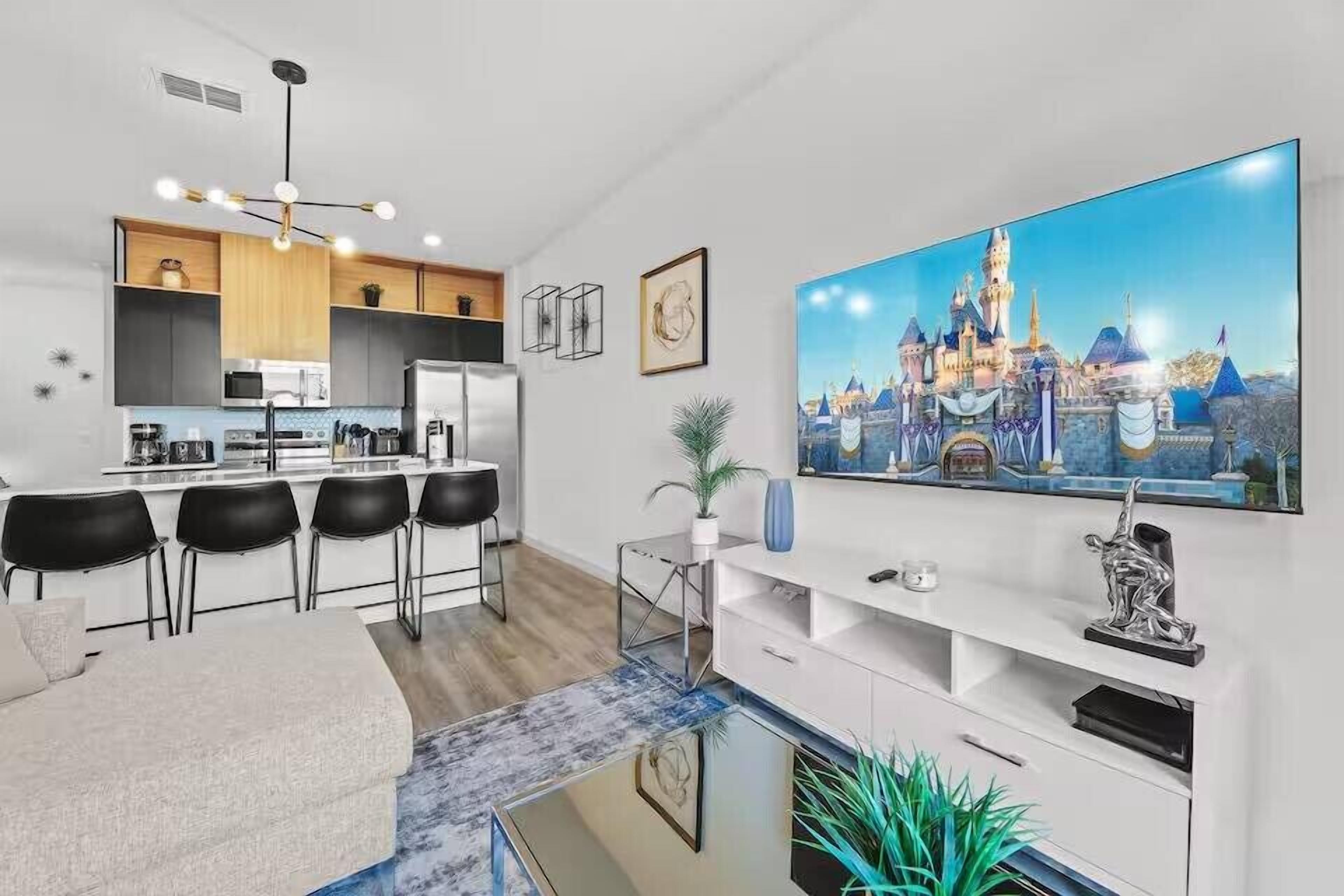 Een moderne keuken met een centraal eiland, een zithoek met een bank en stoelen, een grote flatscreen televisie aan de muur en een decoratieve plant.