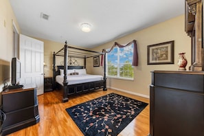 A bedroom with a four-poster bed, a large window with curtains, a dark wooden dresser, and a black floral rug. - Sleeps 6 · 2nd Floor Condo w/ Private Balcony (Orlando)
