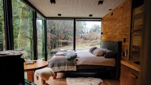 Un dormitorio con un ventanal grande, una cama, suelo de madera y una pequeña mesa redonda.