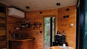 Interior de una cabaña de madera con cama, una mesita, una zona de cocina y vistas a través de un ventanal.