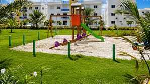 Ein Spielplatz mit Wippe, Klettergerüst und Rutsche, umgeben von einer Rasenfläche und Palmen, mit Gebäuden im Hintergrund.