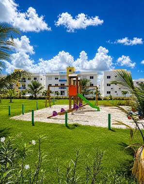 Un parque infantil con sube y baja, estructura para escalar y tobogán, rodeado de césped y palmeras, con edificios al fondo.