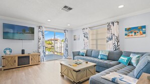 Un salon spacieux avec une grande télévision, une table basse en bois, un canapé gris et un balcon offrant une vue sur une plage et des palmiers.
