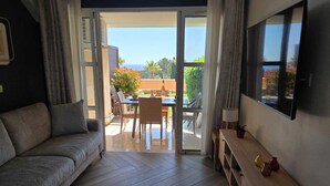 Ein modernes Wohnzimmer mit einer Couch, einem an der Wand montierten Fernseher, einem Essbereich mit Tisch und Stühlen und einer Glasschiebetür, die auf eine Terrasse im Freien führt.