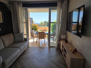 Ein modernes Wohnzimmer mit einer Couch, einem an der Wand montierten Fernseher, einem Essbereich mit Tisch und Stühlen und einer Glasschiebetür, die auf eine Terrasse im Freien führt.