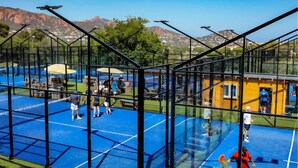 Ein Tennisplatz mit Glasumrandung, umgeben von einem Metallzaun, mit Bergblick im Hintergrund.