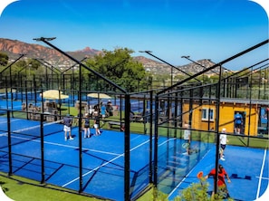 Ein Tennisplatz mit Glasumrandung, umgeben von einem Metallzaun, mit Bergblick im Hintergrund.