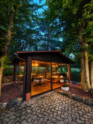 A glass-enclosed outdoor seating area with a wooden table and chairs, surrounded by trees.