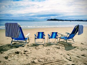 Drei Liegestühle mit Handtüchern an einem Sandstrand, im Hintergrund das Meer.