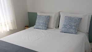 Una cama con ropa de cama blanca y dos almohadas estampadas, una mesita de noche y una ventana con cortinas.