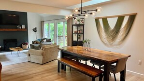 A modern dining area with a wooden table and benches, a black bookshelf, and a large window with a view of greenery. - Woodland Edge Retreat- Borders NRG National Park, trail access from backyard (Fayetteville)