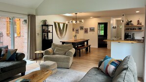 A spacious living area with a sofa, coffee table, and a dining table. There is a kitchen area with a counter and shelves. - Woodland Edge Retreat- Borders NRG National Park, trail access from backyard (Fayetteville)