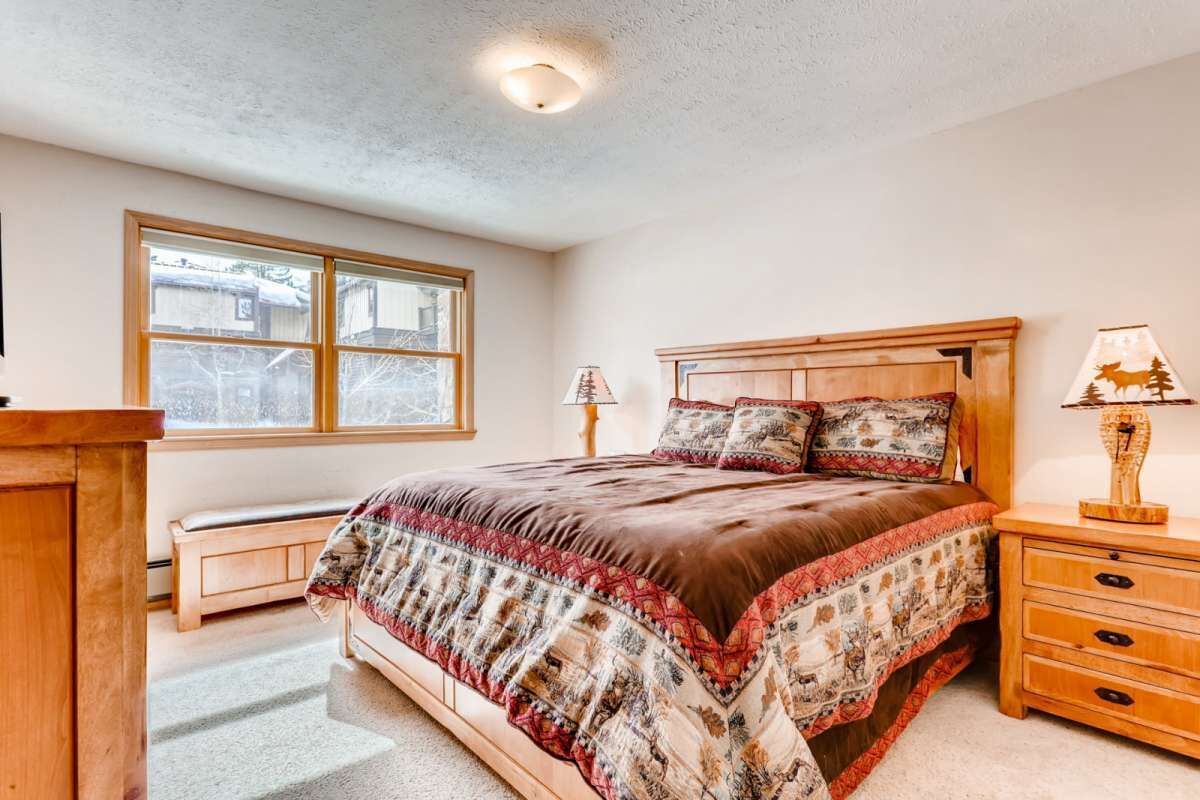 A bedroom with a large bed, a wooden nightstand with lamps, a bench, and a window with a view of a building.