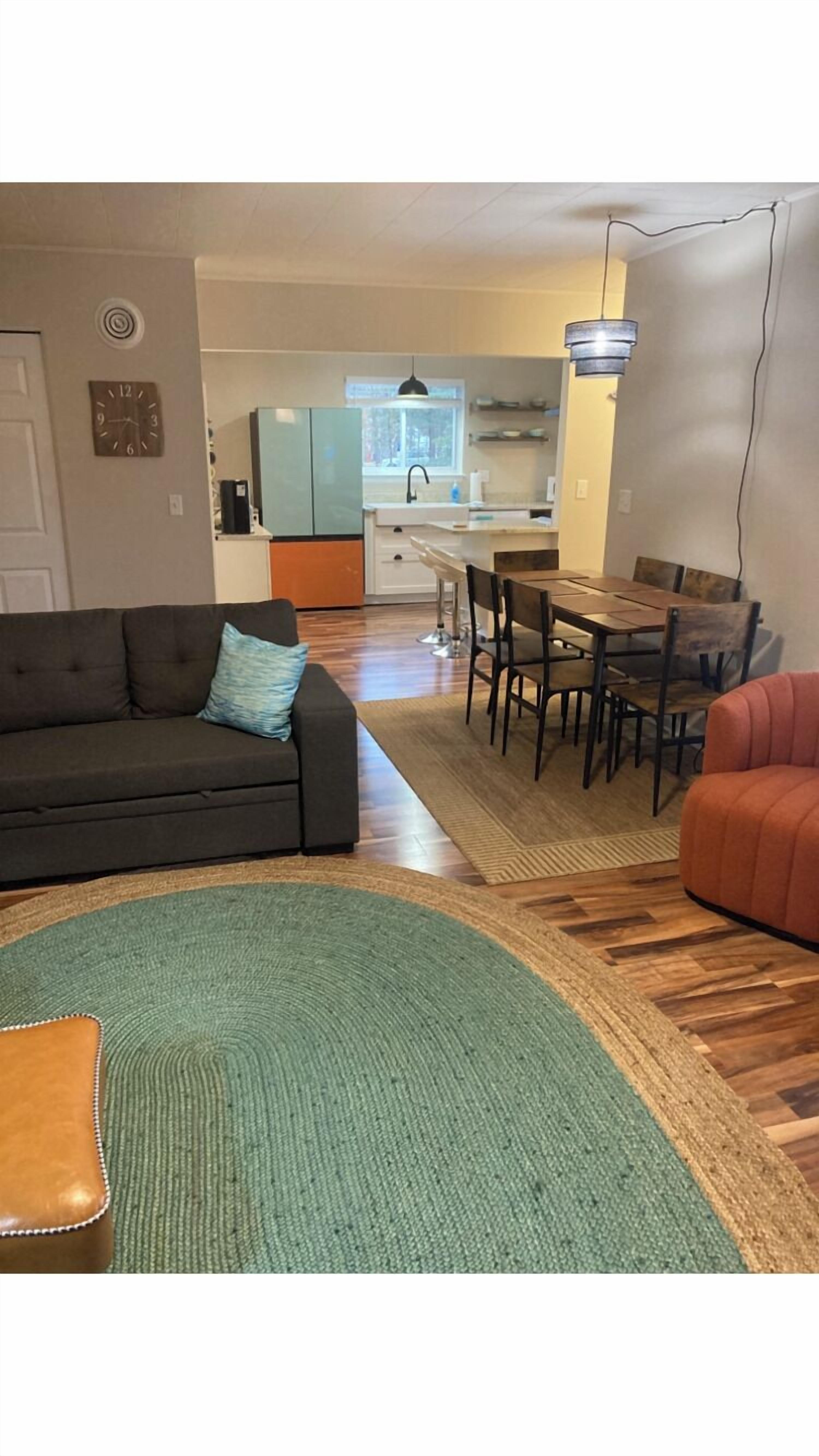 A living room with a sofa, a dining table with chairs, a kitchen area with a window, and a clock on the wall.