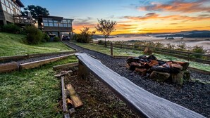 A modern building with a pathway leading to a fire pit area, surrounded by a wooden fence and a view of distant hills. - Chalet Paradise (Bom Jardim da Serra)