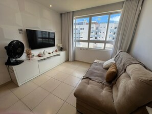 A modern living room with a flat-screen TV, a sofa, a fan, and a window with a view of buildings. - Clocio Beltrão 205 - Beachfront Apartment on Cabo Branco Beach for 4 people (João Pessoa)