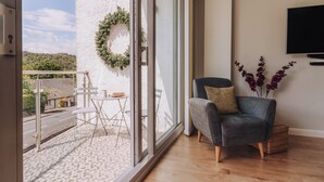 Un balcon avec une balustrade blanche, une petite table avec des chaises, un fauteuil gris avec un coussin, un panier en osier et un téléviseur fixé au mur.