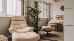 Une pièce avec un grand miroir, un fauteuil beige, une plante en pot, un lit recouvert d’une couverture tissée et un luminaire suspendu.