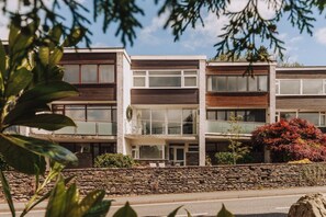 A modern multi-story residential building with a stone wall in front, balconies, and a clear sky. - Nearmere, Slps 8, Central, Parking, Balcony, Views (Bowness-on-Windermere)