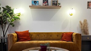 A living room with a round glass coffee table, a mustard-colored sofa with orange cushions, a wooden shelf with framed pictures, and a potted plant. - Flamingo Grove, Vibrant Downtown Studio Escape, AC (Halifax)