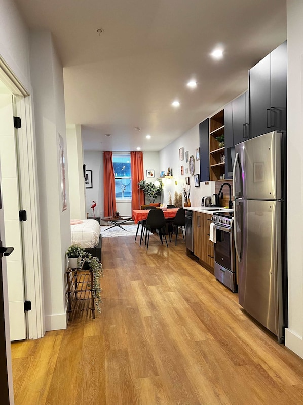 A modern kitchen with wooden flooring, stainless steel appliances, and a dining area with a red tablecloth. - Flamingo Grove, Vibrant Downtown Studio Escape, AC (Halifax)