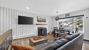 A modern living room with a fireplace, a large TV, and a dining area with a view of the outdoors. - Modern Brian Head Condo - Walk to Giant Steps -  Hot Tub - Pet Friendly (Brian Head)