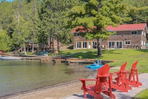 Una cabaña junto al lago con un muelle, sillas Adirondack rojas y un bote.