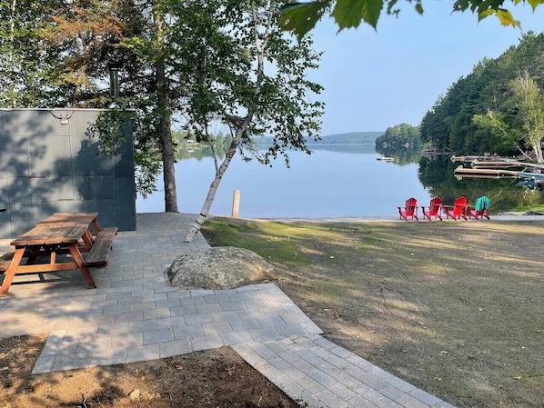 Zona de picnic con banco, paseo pavimentado y sillas rojas junto a un lago.