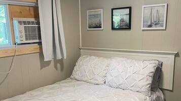 A bed with a quilted white duvet, a white headboard, and a window with an air conditioning unit.