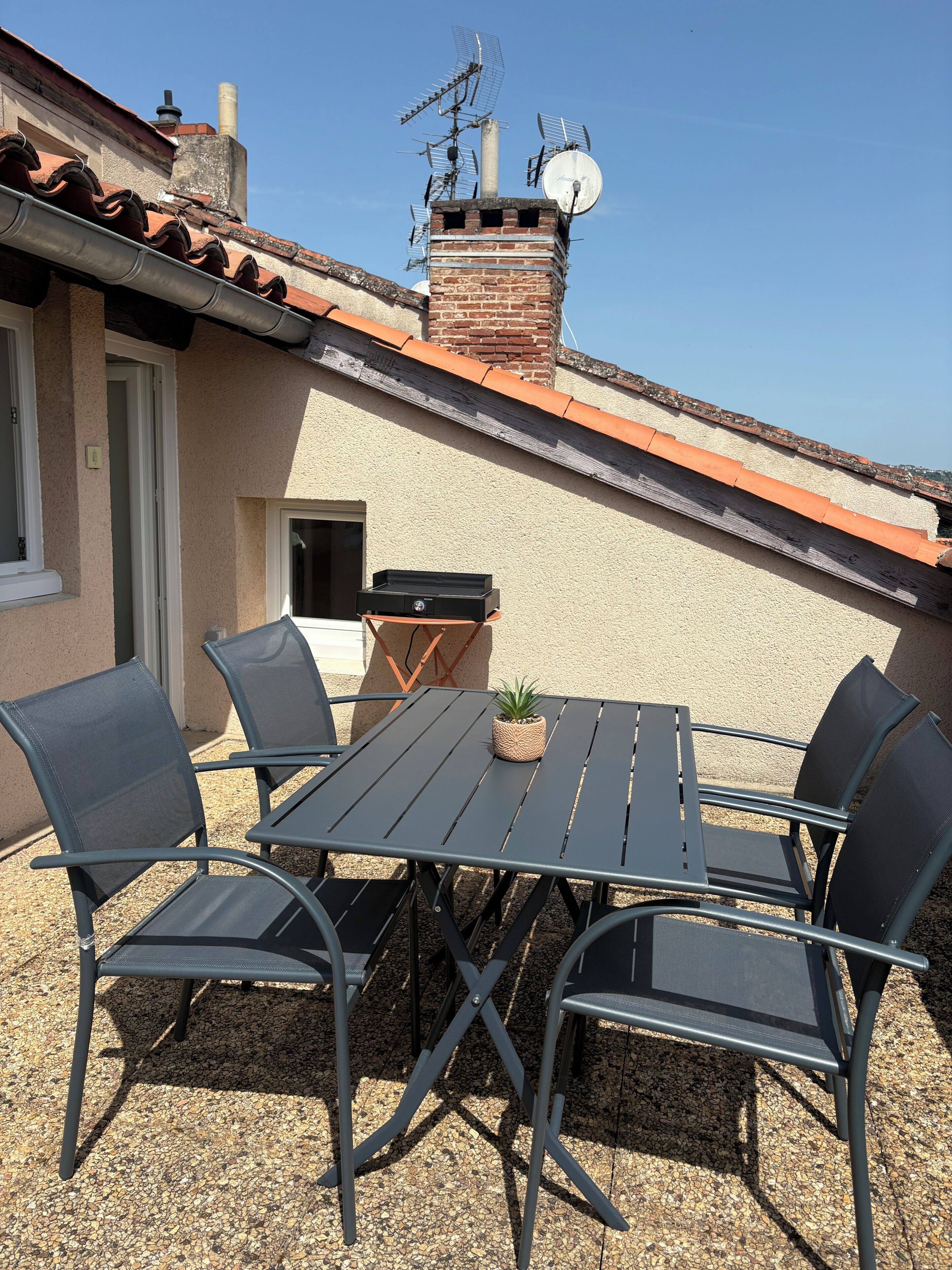 Een terras buiten met een tafel, stoelen, een kleine plant en een schoorsteen op de achtergrond.