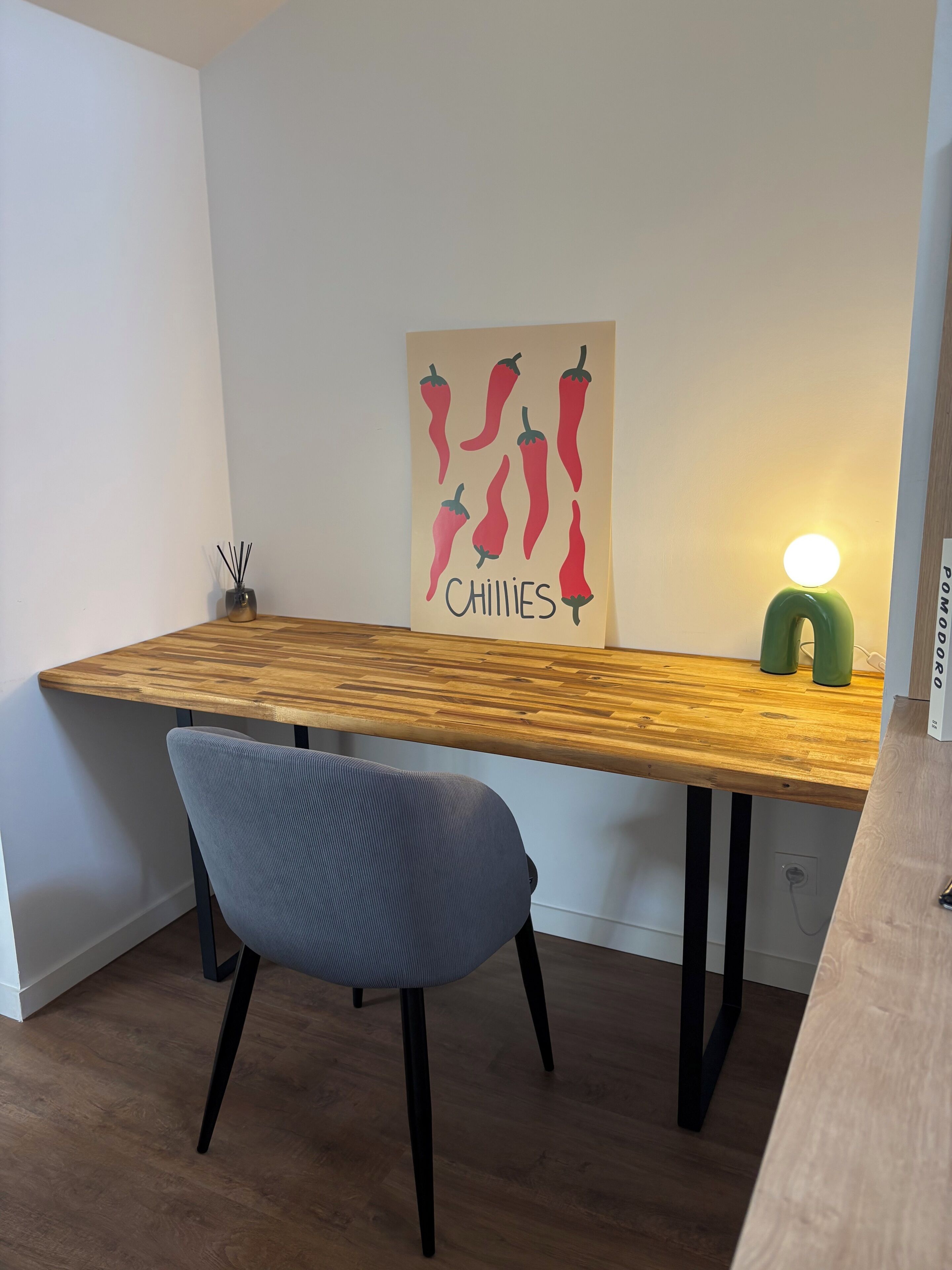 Une table à manger en bois, accompagnée d’une chaise, d’une lampe fixée au mur et d’une affiche représentant des piments.