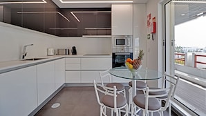 A modern kitchen with white cabinetry, a glass dining table, and a view through a large window. - Marina Luxor 21 in Vilamoura (Loulé)