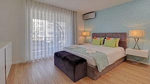 A bedroom with a bed, bedside lamps, a window with curtains, a wall-mounted air conditioner, and a wooden floor. - Marina Luxor 21 in Vilamoura (Loulé)